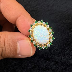 May include: A gold ring featuring a large oval-shaped white opal center stone. The opal is surrounded by a halo of alternating small round clear white stones and small round green stones set in a gold metal frame with a textured gold band.