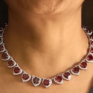 May include: A silver necklace with a heart-shaped pendant design. The pendant is made of red gemstones surrounded by smaller clear gemstones.