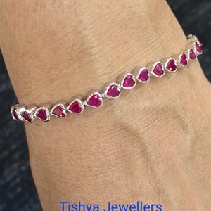 May include: A silver bracelet with heart-shaped, ruby-red gemstones. The bracelet is displayed on a wrist, with the text "Tishya Jewellers" in blue. The gemstones are arranged in a line, creating a delicate and romantic design.