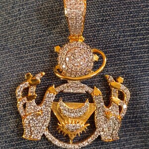 May include: Gold-toned pendant with a detailed design. The pendant features a teapot, hands, and a crescent moon, all encrusted with sparkling crystals. The pendant is set against a denim background.