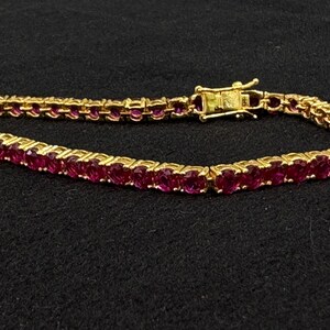 May include: A gold tennis bracelet featuring a continuous row of round, deep pink gemstones set in individual metal prongs. The bracelet includes a secure box clasp mechanism with a safety latch, creating a flexible and circular jewelry piece.