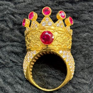 May include: A gold ring featuring a crown design. The crown is adorned with red and white gemstones. This ring is a bold statement piece and would make a wonderful gift.