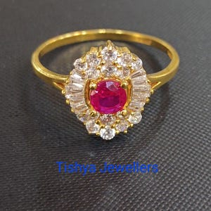 May include: A gold ring with a central, round, ruby-coloured gemstone encircled by smaller, clear gemstones. The ring's band is plain, polished gold. The text "Tishya Jewellers" is at the bottom.