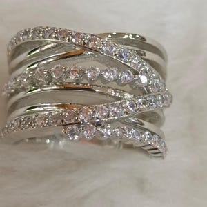 May include: A wide, silver ring with a multi-layered design. The ring features multiple bands that crisscross and overlap, adorned with a row of small, clear gemstones. The ring is set against a soft, white background.