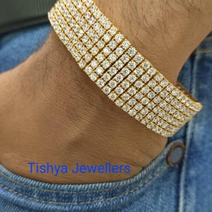 Può includere: Un bracciale d'oro con più file di pietre preziose chiare e scintillanti. Il bracciale è indossato su un polso e il testo "Tishya Jewellers" è visibile sullo sfondo blu denim.