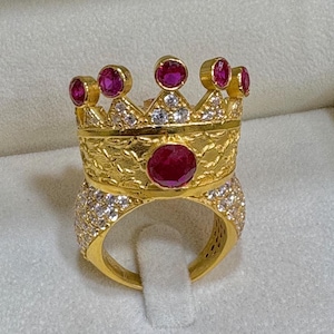 May include: A gold ring with a crown design featuring a large red ruby stone and smaller ruby stones around the crown. The ring is also decorated with small white diamonds.