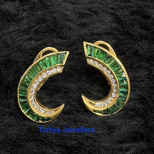 May include: A pair of gold-toned earrings with a curved design. The earrings feature a row of small, sparkling white stones and a border of emerald green stones. The text "Tishya Jewellers" is visible at the bottom.