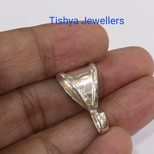 May include: A silver pendant bail with a triangular shape and a polished metallic finish. The side of the bail is clearly stamped with the text 925, indicating sterling silver content. The text Tishya Jewellers is visible above the object.