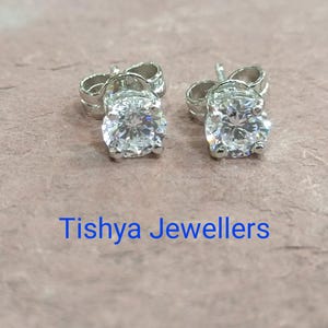 May include: A pair of stud earrings with round, clear gemstones set in silver-colored metal. The earrings are displayed on a textured, light brown surface. The text "Tishya Jewellers" is visible in blue.