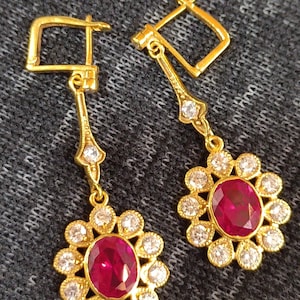 Può includere: Orecchini pendenti color oro con un rubino ovale centrale circondato da un alone di piccole gemme rotonde e trasparenti. Gli orecchini hanno un design floreale e sono sospesi da una chiusura rettangolare. Il testo "Tishya Jewellers" è visibile in basso.