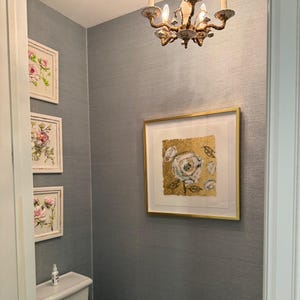 Op de afbeelding: Een kleine badkamer met grijze muren en een wit toilet. Er hangt een goudkleurig ingelijst schilderij met een witte bloem op een gouden achtergrond aan de muur. Er hangen drie kleinere ingelijste schilderijen met bloemmotieven aan de muur links van het grote schilderij.