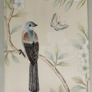 May include: An illustration of a bird perched on a branch, with a butterfly and flowers. The bird has blue, brown, and black feathers. The background is a light beige color, with pink and white flowers.