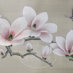 Può includere: Opera d'arte ricamata che presenta un delicato ramo di fiori di magnolia bianchi con riflessi rosa, su uno sfondo beige. Una singola farfalla blu con segni bianchi vola sopra i fiori.