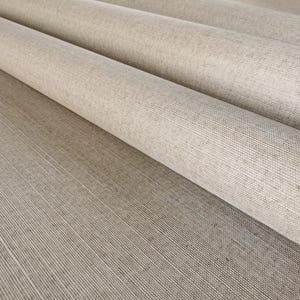 May include: Close-up of rolled beige fabric with a textured surface. The fabric is rolled into several layers, revealing the weave and subtle color variations. The material appears to be a linen or similar textile, suitable for upholstery or drapery.