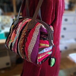 K&ouml;nnte beinhalten: Eine kleine, bunte Patchwork-Handtasche mit einem braunen Lederriemen und einem Rei&szlig;verschluss. Die Tasche ist aus verschiedenen Stoffen gefertigt, darunter Samt, Cord und Baumwolle.
