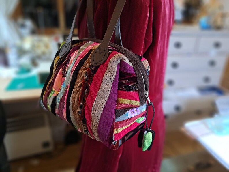 K&ouml;nnte beinhalten: Eine kleine, bunte Patchwork-Handtasche mit einem braunen Lederriemen und einem Rei&szlig;verschluss. Die Tasche ist aus verschiedenen Stoffen gefertigt, darunter Samt, Cord und Baumwolle.