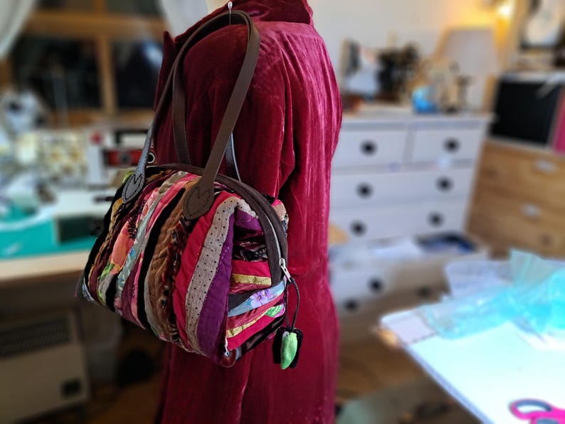 K&ouml;nnte beinhalten: Eine kleine, bunte Patchwork-Handtasche mit braunen Ledergriffen und einem Rei&szlig;verschluss. Die Tasche ist aus verschiedenen Stoffen gefertigt, darunter Streifen, Punkte und Blumenmuster.