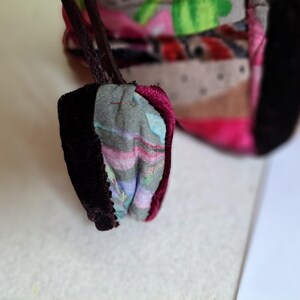 K&ouml;nnte beinhalten: Nahaufnahme einer kleinen, bunten Stofftasche mit dunkelviolettem Samtbesatz. Die Tasche besteht aus Patchwork-Stoff mit verschiedenen Mustern und Farben, darunter Rosa, Gr&uuml;n und Braun.