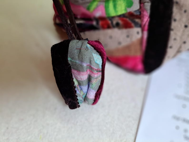 K&ouml;nnte beinhalten: Nahaufnahme einer kleinen, bunten Stofftasche mit dunkelviolettem Samtbesatz. Die Tasche besteht aus Patchwork-Stoff mit verschiedenen Mustern und Farben, darunter Rosa, Gr&uuml;n und Braun.