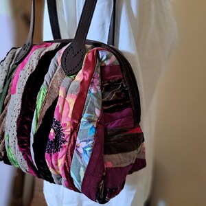 K&ouml;nnte beinhalten: Eine Patchwork-Handtasche mit einem braunen Lederriemen und -griff. Die Tasche ist aus verschiedenen Stoffen in den Farben Rosa, Rot, Braun und Gr&uuml;n gefertigt.