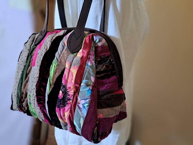 K&ouml;nnte beinhalten: Eine Patchwork-Handtasche mit einem braunen Lederriemen und -griff. Die Tasche ist aus verschiedenen Stoffen in den Farben Rosa, Rot, Braun und Gr&uuml;n gefertigt.