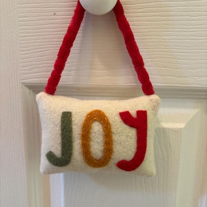 May include: A white felt pillow with the word "JOY" in green, orange, and red letters. The pillow is suspended by a red pipe cleaner and a white wooden ball. The item is hanging on a white door.