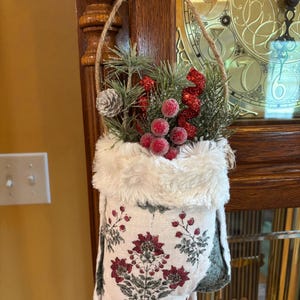 May include: A hanging fabric pouch featuring a white base with a floral pattern in dark red and green. The top edge is trimmed with white faux fur. The pouch is filled with decorative pine branches, a small silver pinecone, and red berries.