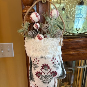 May include: A hanging fabric pouch featuring a white base with a dark red and green floral pattern, topped with a white faux fur cuff. The pouch is filled with decorative pine branches, small pinecones, and three red and white striped spheres.