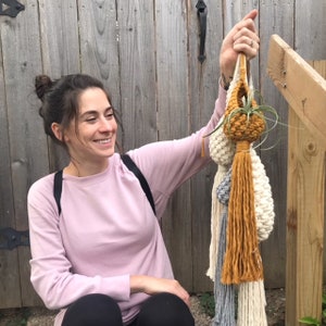 May include: A woman holds up a set of three macrame plant hangers with tassels. The hangers are made with natural fibers in shades of white, cream, and mustard yellow. The top hanger has a small air plant inside.