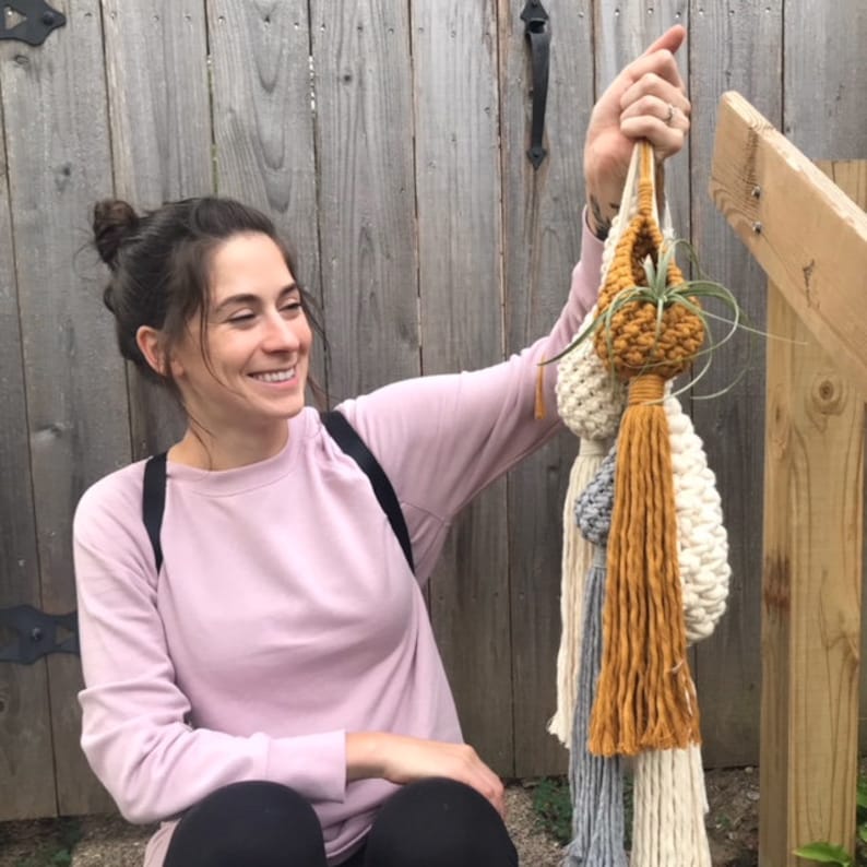 May include: A woman holds up a set of three macrame plant hangers with tassels. The hangers are made with natural fibers in shades of white, cream, and mustard yellow. The top hanger has a small air plant inside.