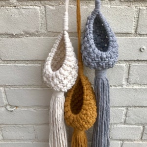 May include: Three macrame plant hangers with different colored pots. The pots are white, mustard yellow, and gray. The hangers are made of natural cotton rope and have long tassels.