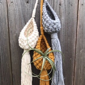 May include: Three macrame plant hangers with different colored pots. The pots are white, mustard yellow, and gray. The hangers are made of natural fiber rope with long tassels.