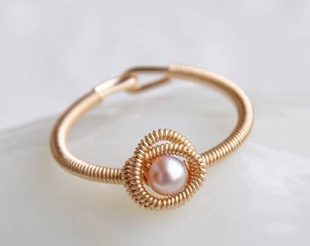 May include: A gold-toned wire-wrapped ring with a central, light pink pearl. The ring's band is a continuous coil, and the pearl is set within a decorative, woven wire setting. The ring is displayed on a white surface.
