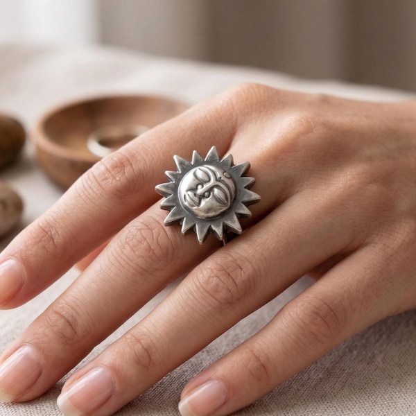 May include: A silver-colored ring featuring a sunburst design with a central face. The ring has a detailed, textured surface and is worn on a finger. The ring is a piece of jewelry.