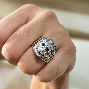 Op de afbeelding: Een zilveren ring met een leeuwenkop en paarse edelsteenogen. De ring wordt gedragen aan een vinger.