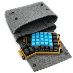 Peut inclure: Un clavier ergonomique divisé avec des touches bleu clair et noires est placé dans un étui de transport en feutre gris. Le clavier a un cadre en bambou. L'étui se ferme avec des boutons-pression noirs.