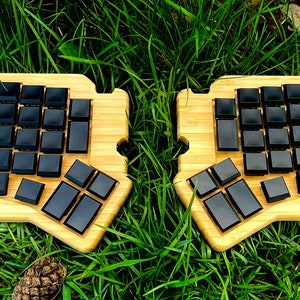 Könnte beinhalten: Geteilte ergonomische Tastatur mit schwarzen Tastenkappen auf einer hellen Bambusbasis. Die Tastatur ist in zwei Hälften geteilt, jede mit einer Reihe von quadratischen und rechteckigen Tasten. Die Tastatur liegt auf einer grünen Wiese.
