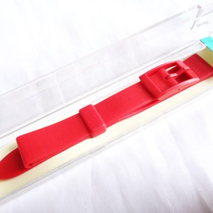 Könnte beinhalten: Ein rotes Uhrenarmband mit einer silbernen Schließe. Das Armband befindet sich in einer durchsichtigen Plastikverpackung mit der Nummer "14" darauf.