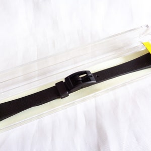 Könnte beinhalten: Ein schwarzes Uhrenarmband mit einer silbernen Schließe. Das Armband befindet sich in einem durchsichtigen Plastiketui mit gelben Etiketten, auf denen "12" steht.