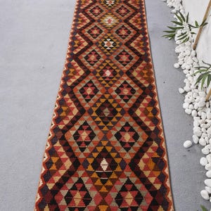 Könnte beinhalten: Ein langer, rechteckiger Läuferteppich mit einem geometrischen Rautenmuster. Der Teppich hat eine Farbpalette aus Rot, Orange, Schwarz und Beige. Das Design umfasst Dreiecke und Rauten, wodurch ein visuell auffälliges Muster entsteht. Der Teppich ist wahrscheinlich aus gewebtem Stoff.