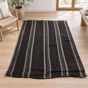 May include: A rectangular area rug featuring a dark charcoal and black base with thin, vertical white stripes running the length of the fabric. The rug has a woven texture and frayed edges along the top and bottom, with a small red tag on the side.