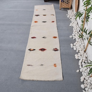 May include: A long, rectangular off-white runner rug featuring a series of small, scattered geometric motifs in shades of red, orange, and black. The textile has a flat-woven texture and finished edges, displaying a repeating pattern of diamond-shaped symbols arranged in rows along the length of the fabric.