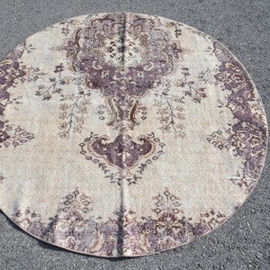May include: A round rug with an ornate, vintage-style design. The rug features a central medallion and border in shades of purple and brown against a cream background. The rug is placed on a gray surface.