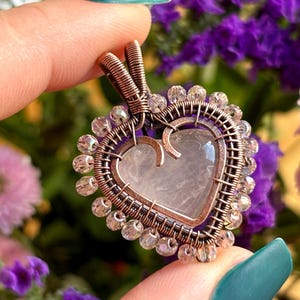 May include: A heart-shaped pendant with a rose quartz centre, encased in copper wire. The pendant is adorned with clear, faceted beads around the perimeter. The background features a blurred arrangement of purple and pink flowers.