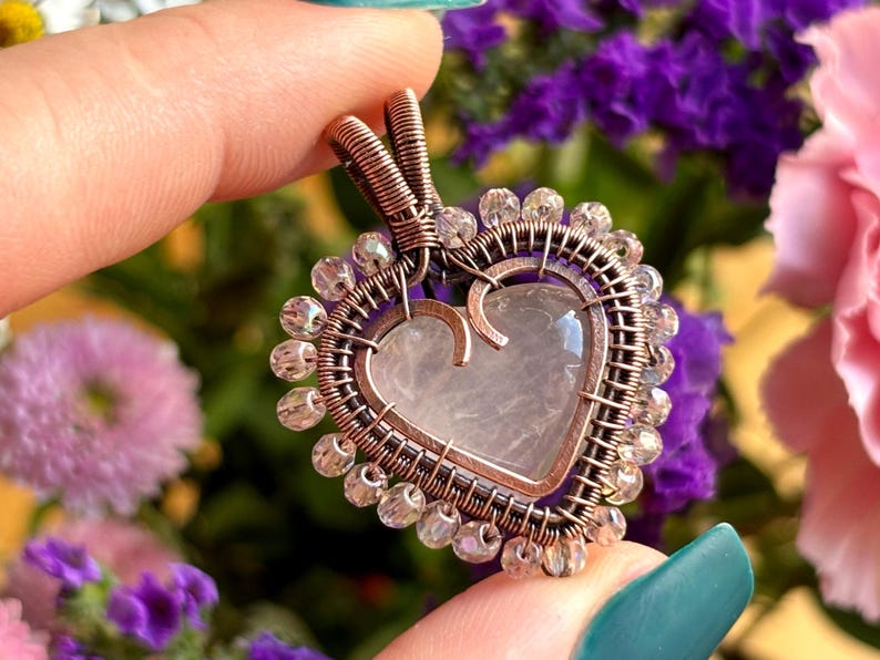 May include: A heart-shaped pendant with a rose quartz centre, encased in copper wire. The pendant is adorned with clear, faceted beads around the perimeter. The background features a blurred arrangement of purple and pink flowers.