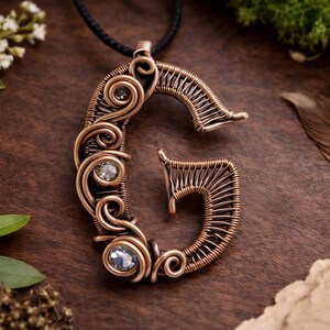 Może przedstawiać: Miedziany wisiorek z drutu w kształcie litery "G", z trzema przezroczystymi, okrągłymi kamieniami szlachetnymi. Wisiorek wisi na czarnym sznurku. Projekt charakteryzuje się skomplikowanymi wirami i teksturowanym wykończeniem na ciemnym tle drewnianym.