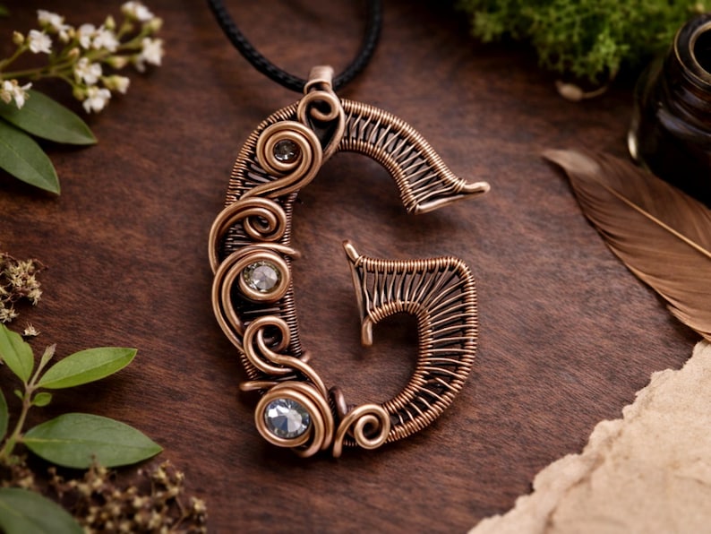 Może przedstawiać: Miedziany wisiorek z drutu w kształcie litery "G", z trzema przezroczystymi, okrągłymi kamieniami szlachetnymi. Wisiorek wisi na czarnym sznurku. Projekt charakteryzuje się skomplikowanymi wirami i teksturowanym wykończeniem na ciemnym tle drewnianym.