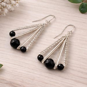 K&ouml;nnte beinhalten: Ein Paar silberfarbene Ohrringe mit geometrischem Design. Jeder Ohrring hat drei silberne St&auml;be und drei schwarze, facettierte Perlen. Die Ohrringe haben einen Hakenverschluss und werden auf einer hellen Holzoberfl&auml;che pr&auml;sentiert.