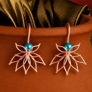 May include: A pair of copper-colored wire earrings shaped like lotus flowers, each featuring a single bright turquoise bead at the center. The earrings have curved wire hooks for attachment and are displayed against a smooth, curved terracotta surface.