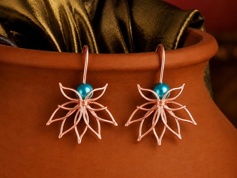 May include: A pair of copper-colored wire earrings shaped like lotus flowers, each featuring a single bright turquoise bead at the center. The earrings have curved wire hooks for attachment and are displayed against a smooth, curved terracotta surface.